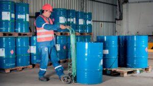 Trabalhador com colete e capacete movimenta tambores de líquido inflamável em galpão de armazenamento de produtos químicos